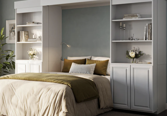 A pull-down white Murphy bed with warm earth-toned pillows and bedding, framed by matching built-in bookcases and storage cabinets.